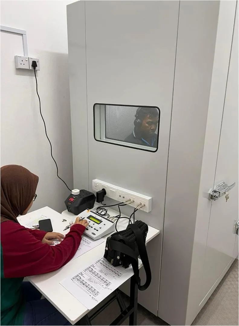 Booth audiometri kalis bunyi dengan juruteknik di luar dan subjek di dalam booth memakai fon kepala.
